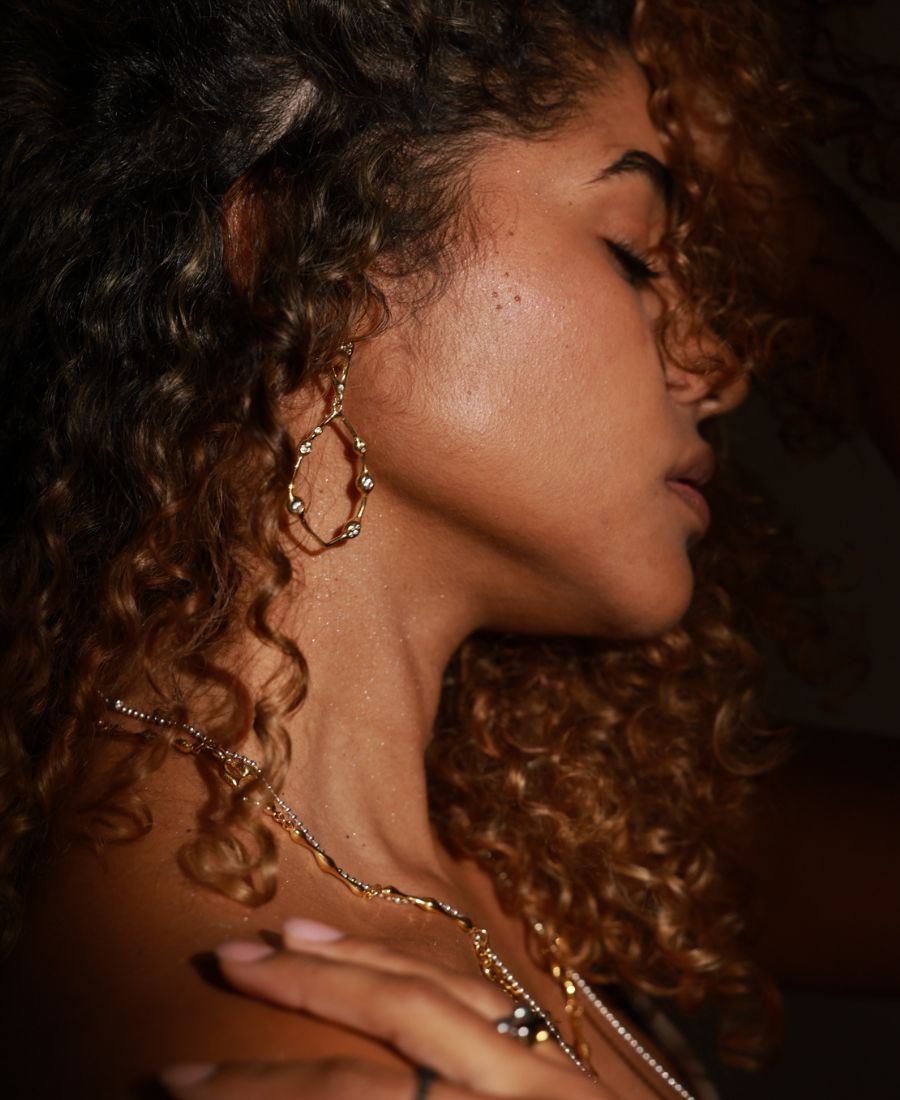 Close-up of a woman with curly hair wearing gold jewelry JYOCO.