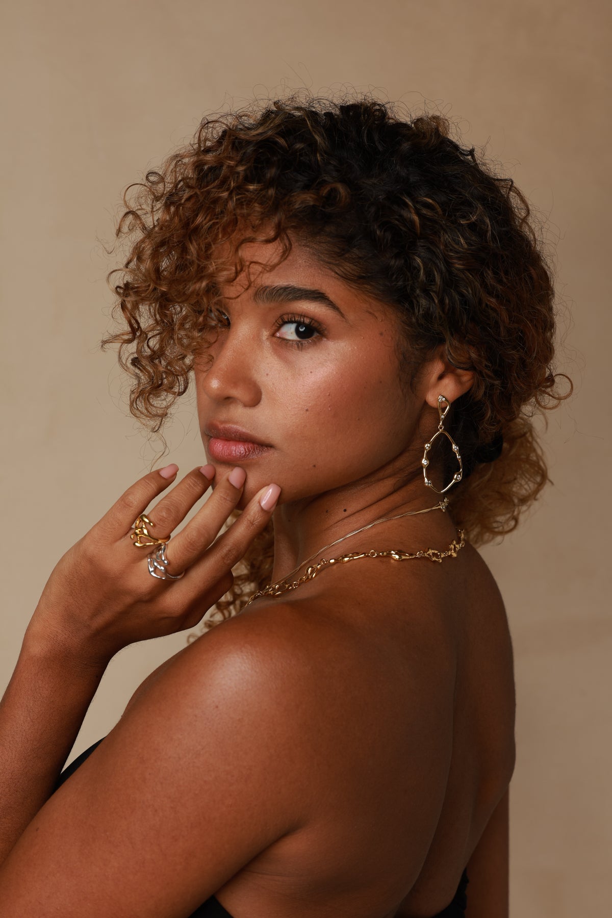 Woman with curly hair wearing JYOCO gold jewelry against a beige background